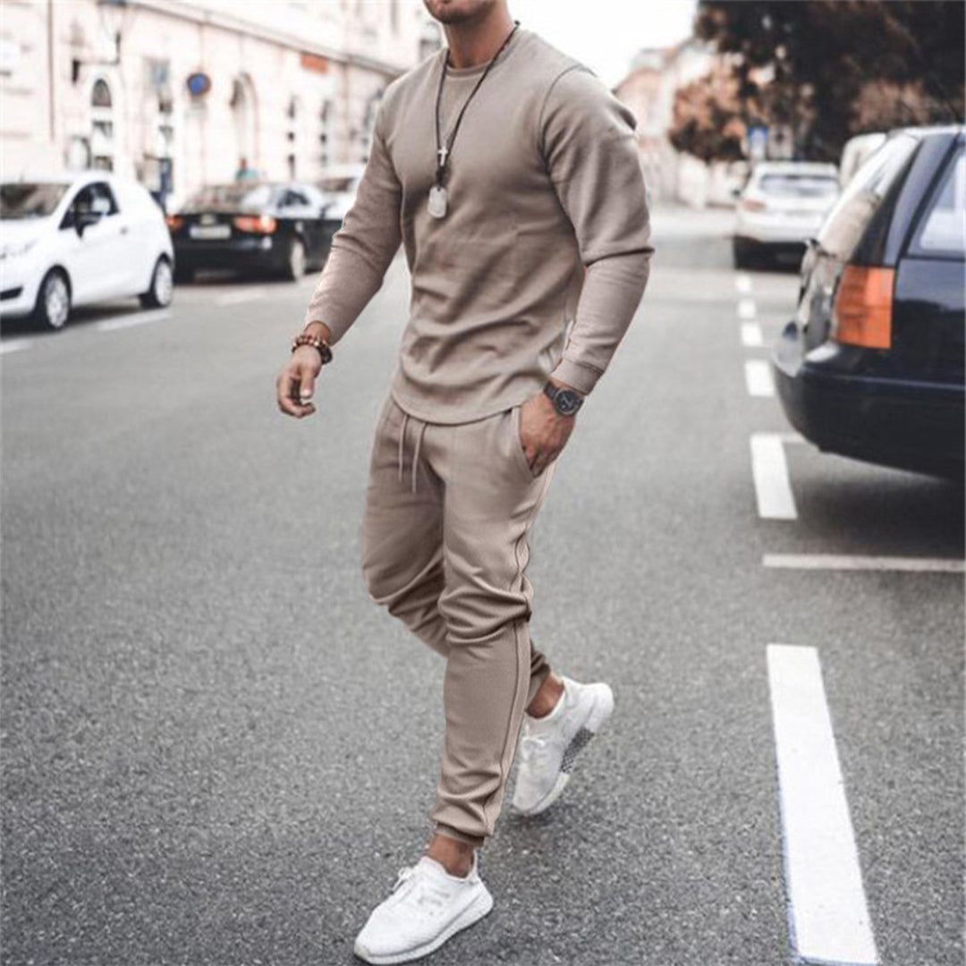 Man in beige trainingspak loopt op straat, draagt witte sneakers en horloge. Stijlvolle herenkleding, casual mode, stedelijke achtergrond.